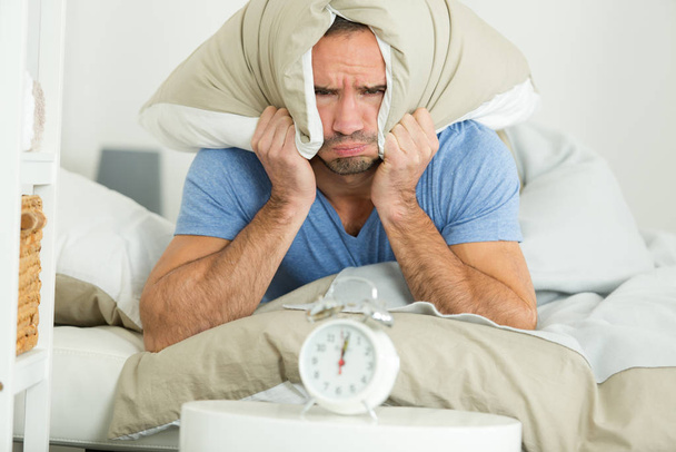 man covering his ears with pillow looking at alarm clock - Foto, imagen