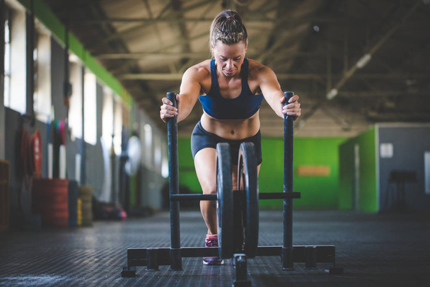 Female fitness model pushing a sled in a cross fit gym. - 写真・画像