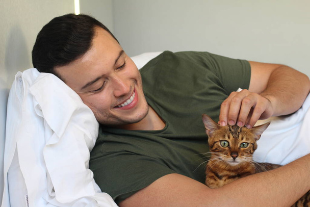 κοντινό πορτραίτο ενός όμορφου νεαρού άντρα που χαλαρώνει στο κρεβάτι με τη γάτα του στο σπίτι - Φωτογραφία, εικόνα