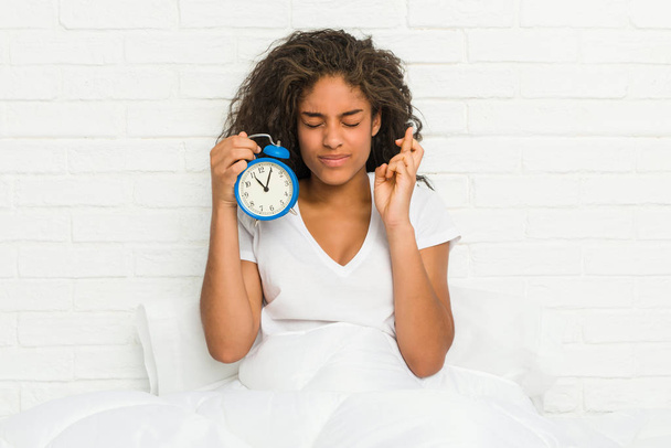 Young african american woman sitting on the bed holding an alarm clock crossing fingers for having luck - Fotografie, Obrázek