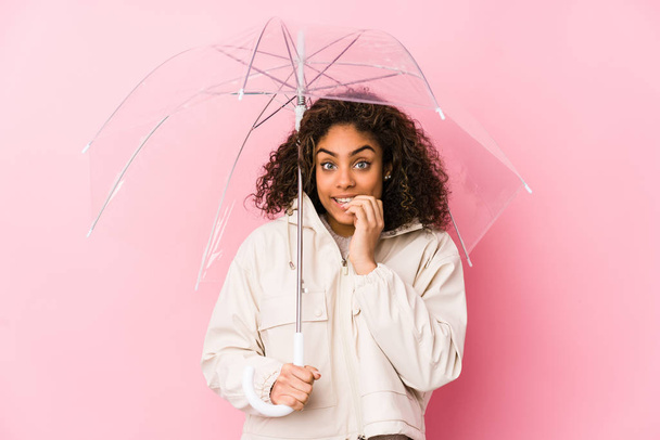 Joven mujer afroamericana sosteniendo un paraguas mordiendo las uñas, nerviosa y muy ansiosa
. - Foto, Imagen