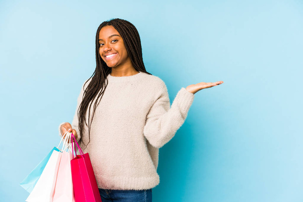 Young african american woman holding a shopping bag isolated showing a copy space on a palm and holding another hand on waist. - Photo, Image