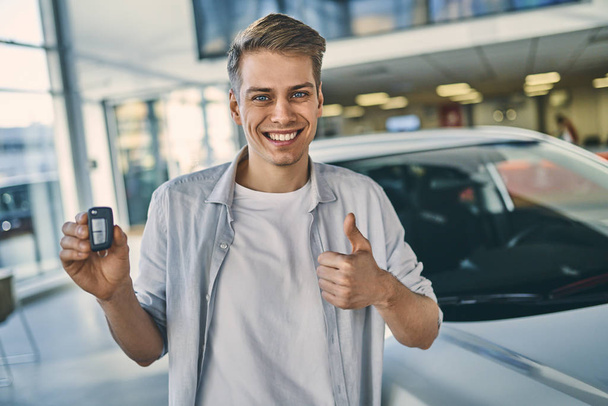 Sonriente hombre sosteniendo una llave detrás de un coche en la sala de exposición. Empresarios
 - Foto, imagen