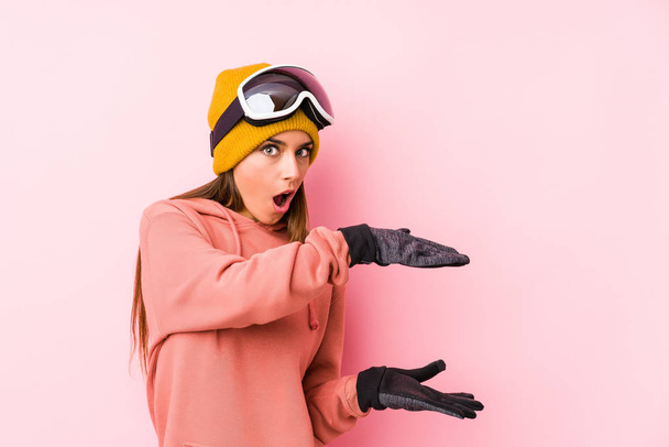 Νεαρή Καυκάσια γυναίκα φορώντας ρούχα του σκι απομονωμένη σοκαρισμένη και έκπληκτη κρατώντας ένα αντίγραφο χώρου ανάμεσα στα χέρια. - Φωτογραφία, εικόνα