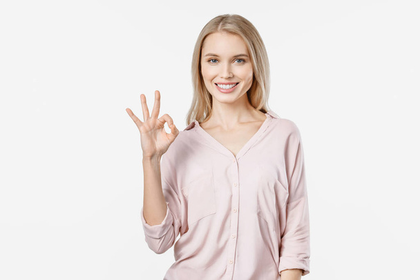Young blonde woman in casual showing OK gesture and looking at camera isolated on white background - Фото, изображение