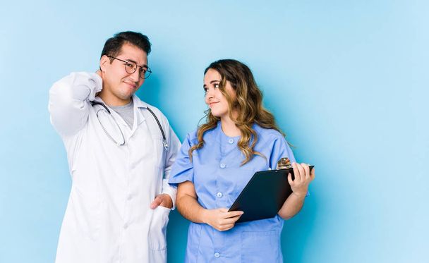 Young doctor couple posing in a blue background isolated suffering neck pain due to sedentary lifestyle. - 写真・画像