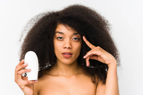 junge Afro-Frau hält eine isolierte Feuchtigkeitscreme und zeigt mit dem Finger auf seine Schläfe, denkt nach, konzentriert sich auf eine Aufgabe. - Foto, Bild