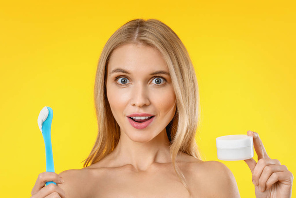 Surprised beautiful woman holding skin cream over yellow background - Fotó, kép