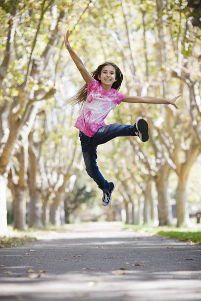 Portrait of Smiling Tween Girl Jumping on Pathway between Trees - Foto, immagini