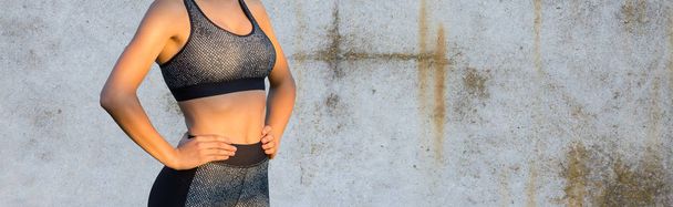 Een sportief slank meisje in legging en een top staat tegen een betonnen muur en doet een set oefeningen. - Foto, afbeelding