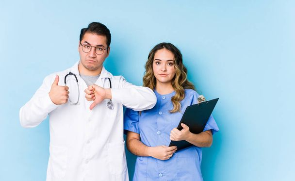 Молода пара лікарів позує на синьому фоні ізольовані, показуючи великі пальці вгору і великі пальці вниз, важко вибрати концепцію
 - Фото, зображення