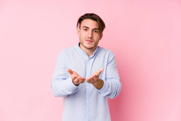 Young caucasian man posing in a pink background isolated holding something with palms, offering to camera. - Foto, immagini