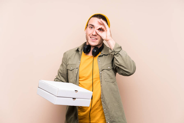 Jovem caucasiano homem segurando pizzas animado mantendo ok gesto no olho
. - Foto, Imagem