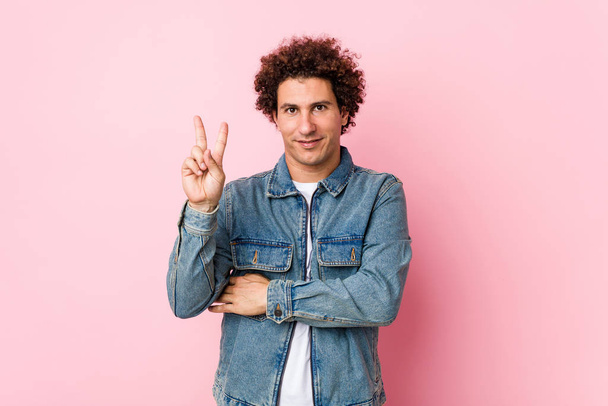 Kudrnatý dospělý muž v džínové bundě na růžovém pozadí ukazující číslo dvě s prsty. - Fotografie, Obrázek