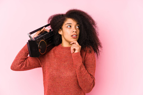 junge Afro-Frau hält eine Kassette isoliert zur Seite schauend mit zweifelndem und skeptischem Gesichtsausdruck. - Foto, Bild