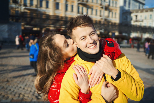 outdoor portrait of young happy couple in love spending time together in the city. woman kissing in cheek her smilling boyfriend. - Фото, изображение