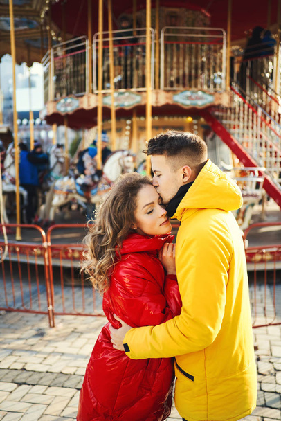 young romantic couple in love wearing in bright yellow and red down jackets hugging on french carousel background. - Photo, Image