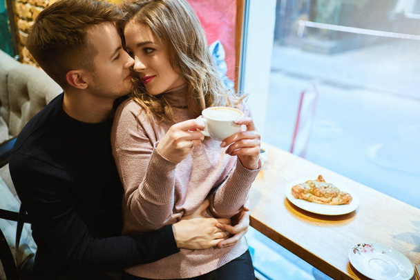 Happy romantic couple sitting over the window in cafe, embracing, kissing and drinking coffee. boyfriend tenderly hugging girlfriend in cozy cafe. - Zdjęcie, obraz