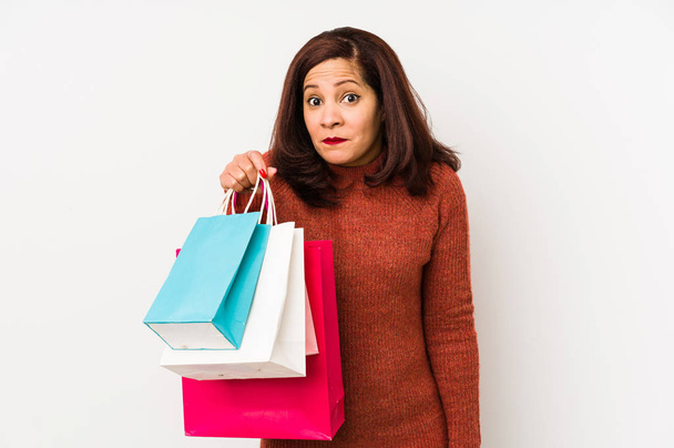 Middle age latin woman holding a shopping bags isolated shrugs shoulders and open eyes confused. - Photo, Image