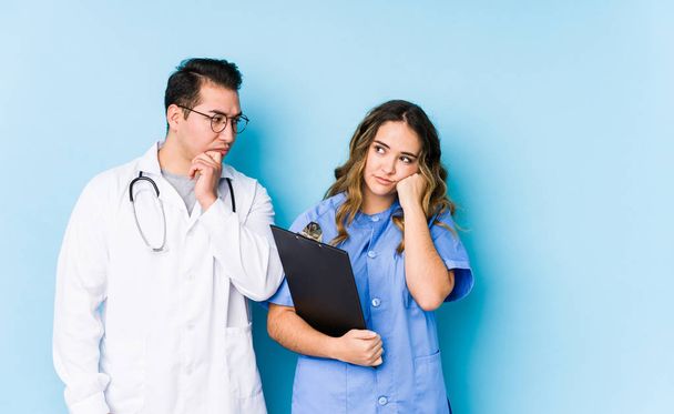 Giovane coppia medico posa in uno sfondo blu isolato che si sente triste e pensieroso, guardando lo spazio copia
. - Foto, immagini