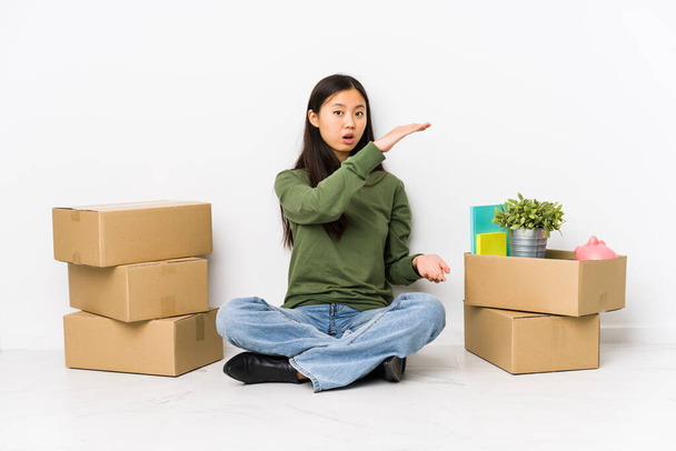 Joven mujer china mudándose a un nuevo hogar sorprendido y sorprendido sosteniendo un espacio de copia entre las manos
. - Foto, Imagen