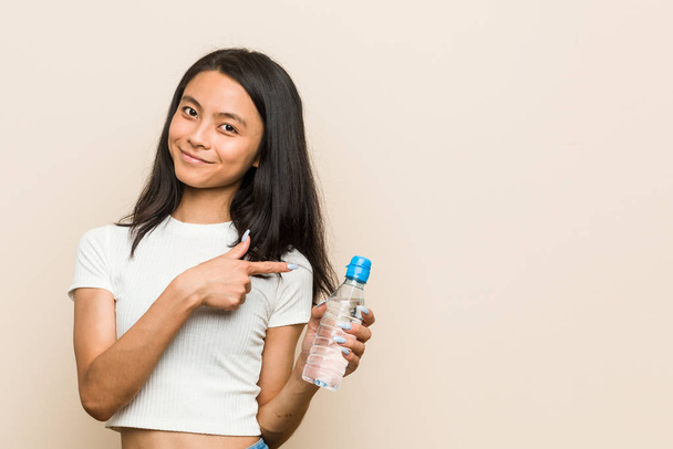 Young asian woman holding a water bottle smiling and pointing aside, showing something at blank space. - Foto, Imagem