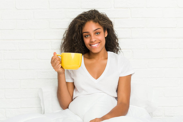 Fiatal afro-amerikai nő ül az ágyon kezében egy kávésbögre boldog, mosolygós és vidám. - Fotó, kép