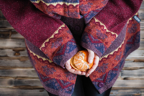 Woman in a huge winter sweater sits on the wooden rustic floor and cleaning the mandarin - Foto, afbeelding