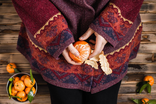 Woman in a huge winter sweater sits on the wooden rustic floor and cleaning the mandarin - Photo, image
