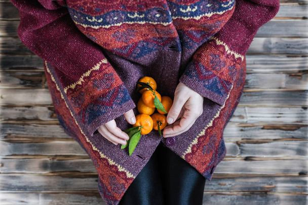 Woman in a huge winter sweater sits on the wooden rustic floor with small iron bucket of tangerines - Photo, image