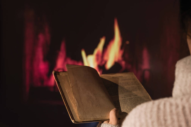 Woman in a winter sweater reading the book sitting near Christmas fireplace with a cup of hot drink and warming woolen blanket. Winter and Christmas holidays concept - Photo, image