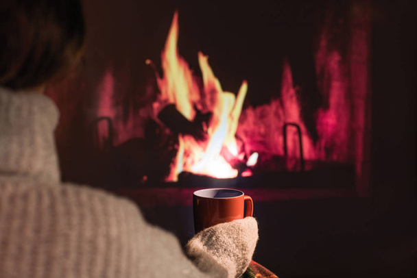 Woman in a winter sweater relaxes sitting near Christmas fireplace with a cup of hot drink and warming woolen blanket. Cozy atmosphere. Winter and Christmas holidays concept - Photo, Image