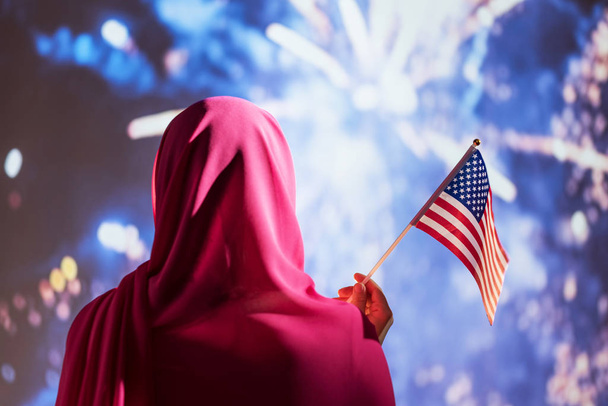 Muslim woman in a scarf holding American flag  during fireworks at night.  - Valokuva, kuva