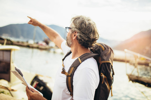 Улыбающийся взрослый турист, стоящий рядом с картой и рюкзаком
 - Фото, изображение