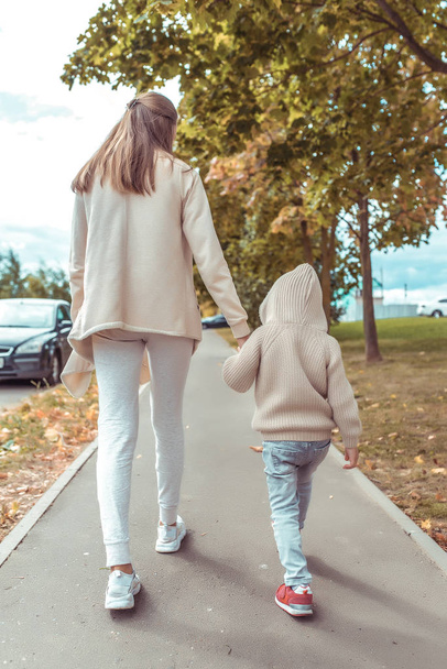 Woman mother walks with her child, little son boy 4-5 years old, autumn day on the street in the city, background road and trees, casual warm clothes with a hood. - Фото, изображение