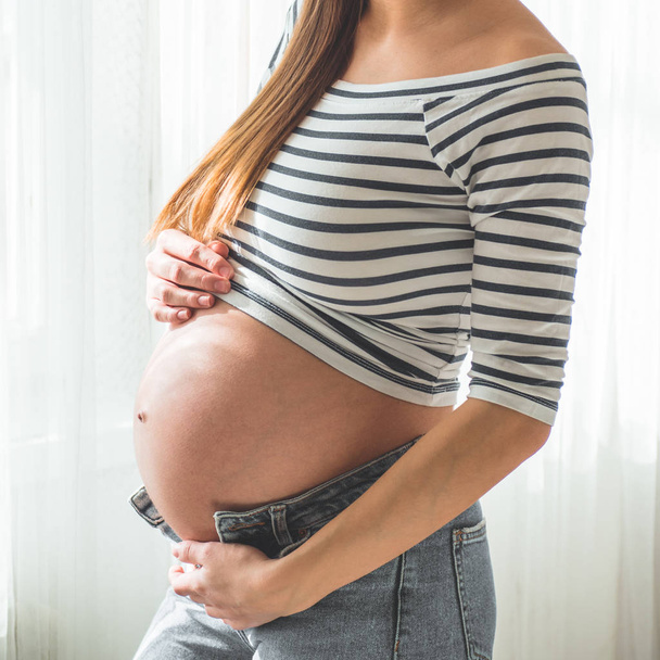Šťastná těhotná žena s velkým břichem u okna. Koncepce těhotenství a rodiny - Fotografie, Obrázek