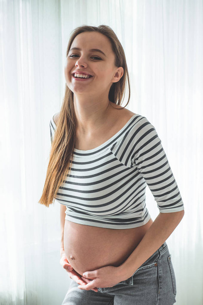 Onnellinen raskaana oleva nainen, jolla on iso vatsa ikkunan vieressä. Raskauden ja perheen käsitteet
 - Valokuva, kuva