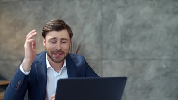 Улыбающийся бизнесмен чатится онлайн на ноутбуке с камерой в офисе. Парень машет рукой
. - Кадры, видео
