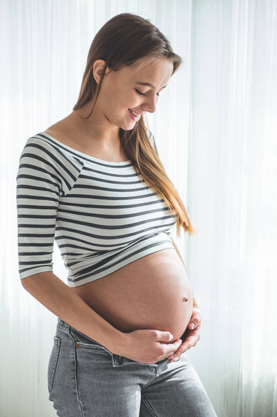 Onnellinen raskaana oleva nainen, jolla on iso vatsa ikkunan vieressä. Raskauden ja perheen käsitteet
 - Valokuva, kuva