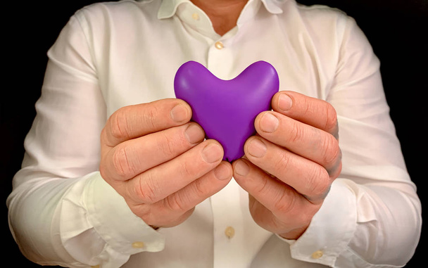 Mies pitää violettia sydäntä käsissään. Mies valkoisessa paidassa, jolla on sydänmerkki, mustalla taustalla. Käsite: suojella, vartija, rakkaus
. - Valokuva, kuva