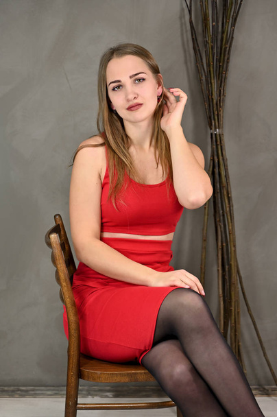 Vertical photo of a pretty smiling girl with long hair and excellent make-up in a red dress sitting on a chair in various poses. Universal concept female portrait on a gray background. - Valokuva, kuva