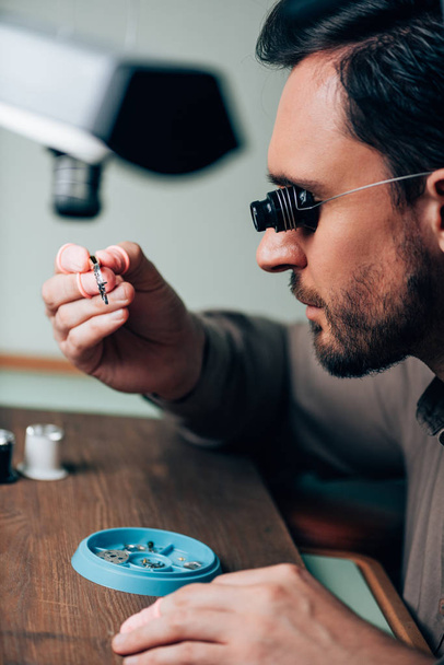 Πλευρική όψη ωρολογοποιού σε εργαλείο γυαλιών που κρατά το ρολόι σε τσιμπίδες στο τραπέζι - Φωτογραφία, εικόνα