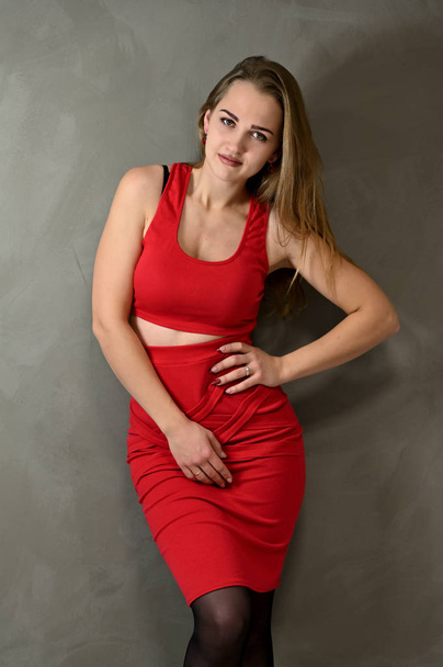 Vertical Photo of a pretty smiling girl with long hair and excellent make-up in a red dress stands next to the wall in various poses. Universal concept female portrait on a gray background. - Photo, image