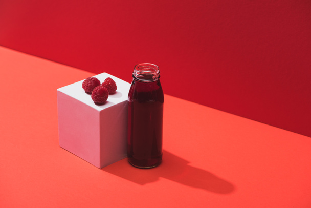 fresh berry juice in glass bottle near ripe raspberries on cube on red background - Photo, Image