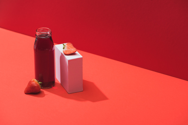 frischer Beerensaft in Glasflasche in der Nähe reifer Erdbeeren auf Würfel auf rotem Hintergrund - Foto, Bild