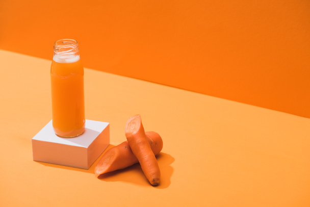 fresh juice in glass bottle on cube near ripe carrots on orange background - Фото, изображение