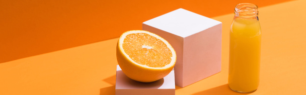 オレンジの半分近くのグラスボトルとオレンジの背景の白い立方体のフレッシュジュースパノラマショット - 写真・画像