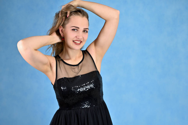 A photo of a pretty smiling girl with long hair and excellent make-up in a black dress stands in different poses. Universal concept horizontal portrait of a woman on a blue background. - Fotografie, Obrázek