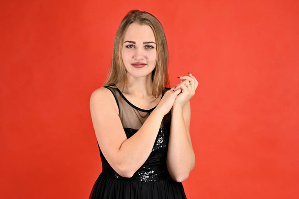 A photo of a pretty smiling girl with long hair and excellent make-up in a black dress stands in different poses. Universal concept horizontal female portrait on a red background. - Foto, Imagem