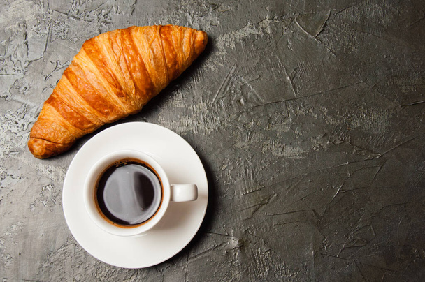 Koffie in witte beker en croissant op een donkere betonnen achtergrond, bovenaanzicht, vlakke lay. Concept voor ontbijt, koffiepauze of zakenlunch. Er is rechts een plaats voor tekst. - Foto, afbeelding